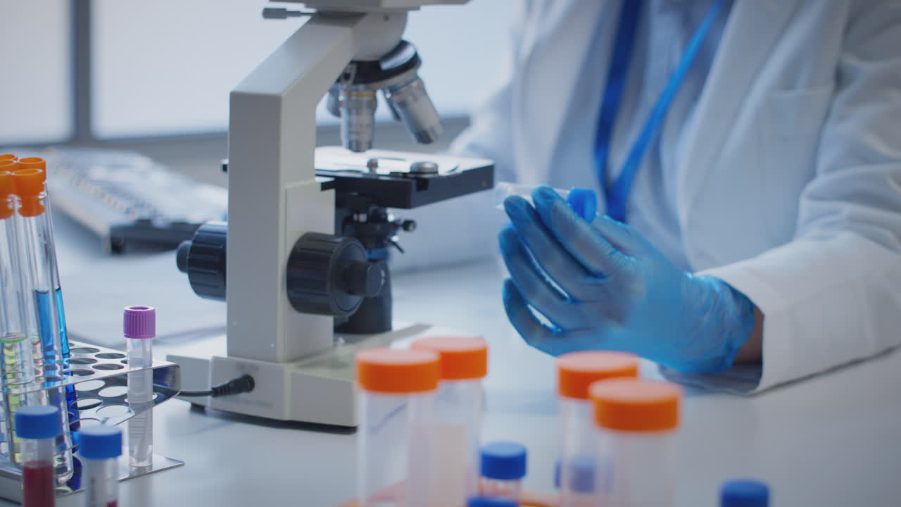 primer plano de un trabajador de laboratorio que realiza investigaciones utilizando un microscopio sosteniendo un tubo de ensayo etiquetado eta
