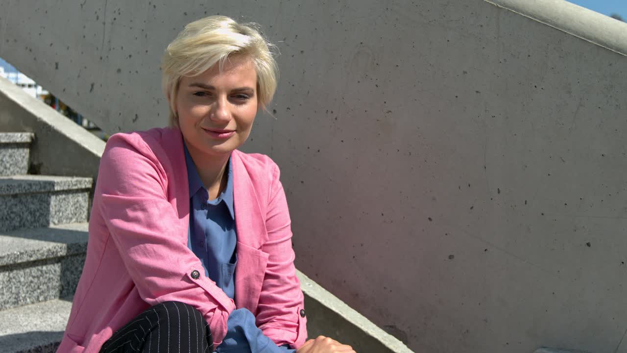 Primo piano di una bella donna che sorride guardando la telecamera, indossando il suo blazer rosa mentre è seduta sulle scale.
