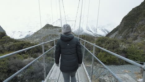 a following shot from behind of a woman walking across a swing bridge over a running stream surrounded by snow capped mountains on a cold winters day in new zealand