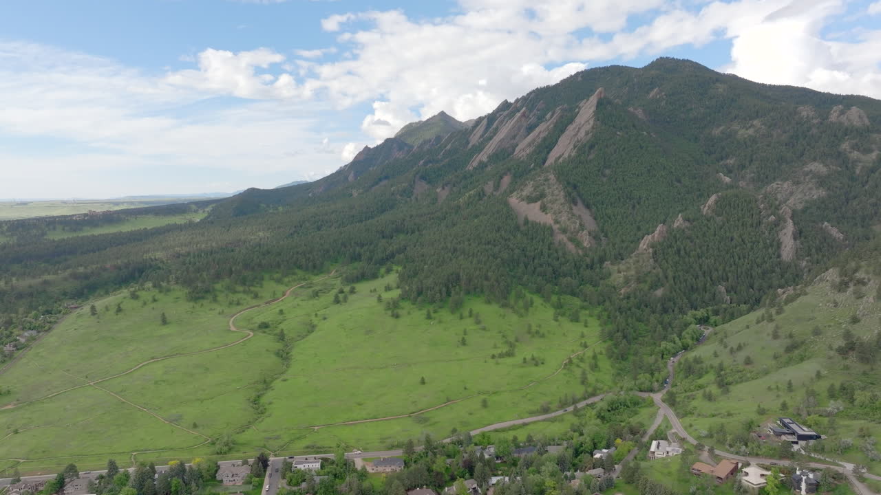 Aerial descend left shot of warm sun hitting Boulder Colorado Flatiron mountains above Chautauqua Park with full green pine trees and blue skies with clouds on a beautiful summer day for hiking