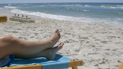 las piernas y los pies de una chica bronceados en el sol frente al océano y un castillo de arena