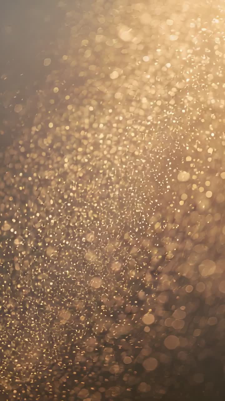 Video verticale: goccioline scintillanti che fluttuano con un leggero sfocato, luce solare dall'alto a destra che crea un bokeh caldo.