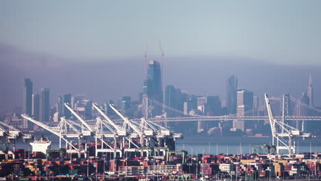 cranes in the san francisco bay loading and unloading cargo ships with the skyline in the foggy background - time lapse