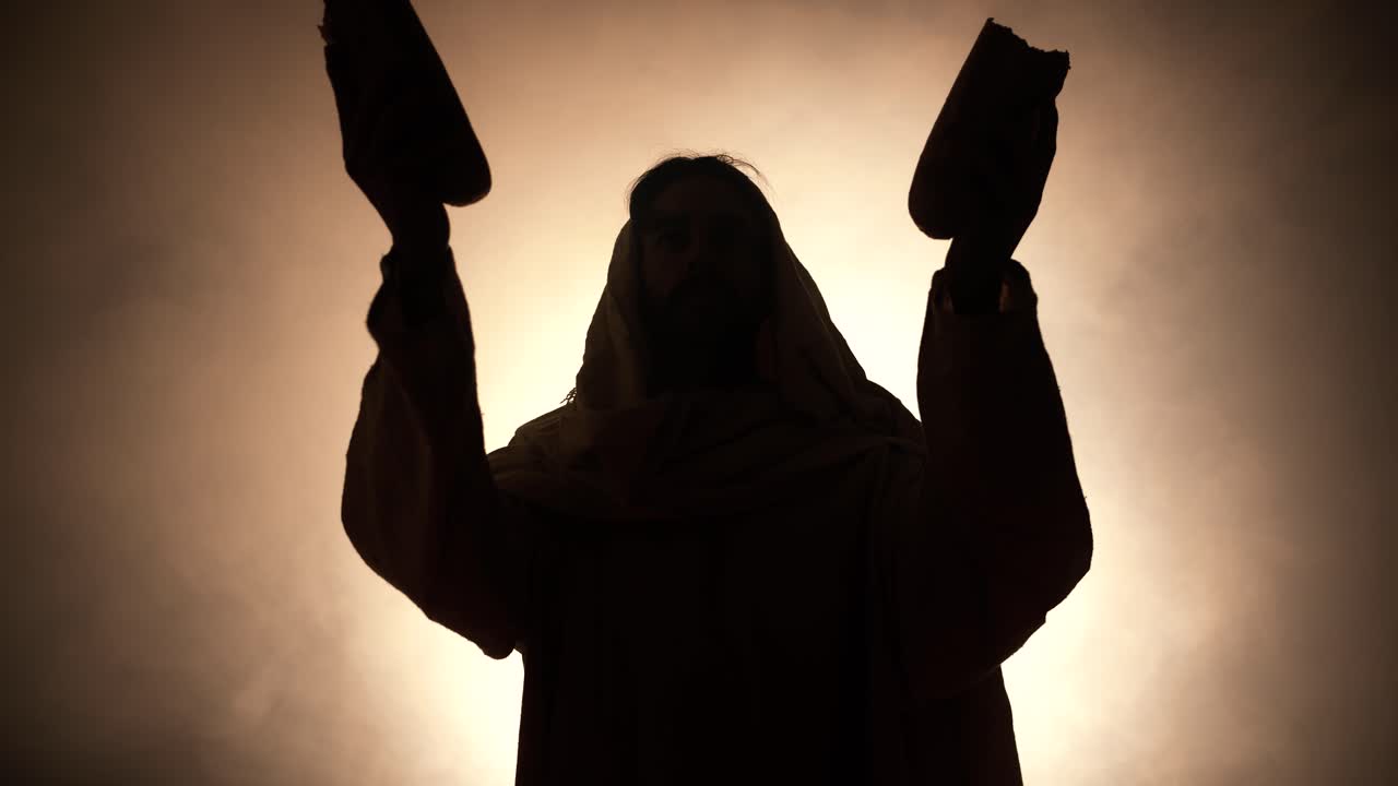 Silhouette of Jesus Christ in white hooded robe holding bread as he stands in glowing, bright light praying. Perfect for representation of Jesus as the bread of life, last supper, communion.