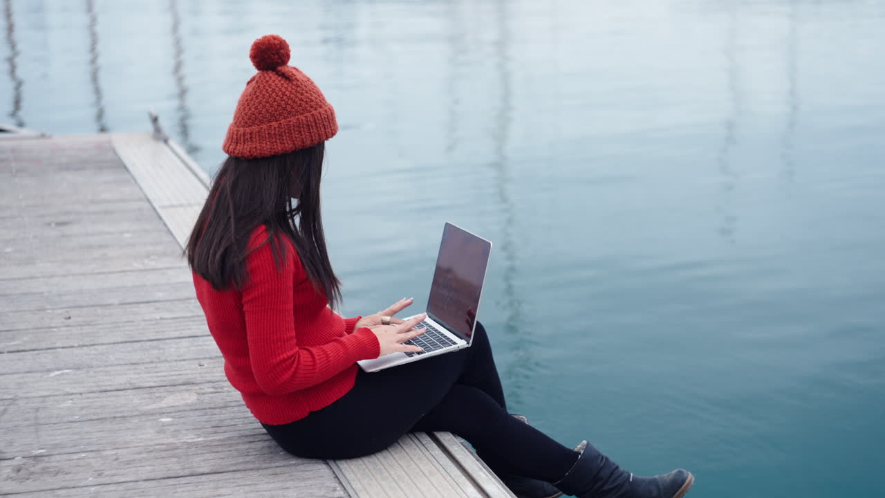 Asian woman sits by the water at the port of Valencia ES, engrossed in her work on a laptop, her thoughts consumed by the task at hand, wearing a red beanie that complements her red turtleneck shirt