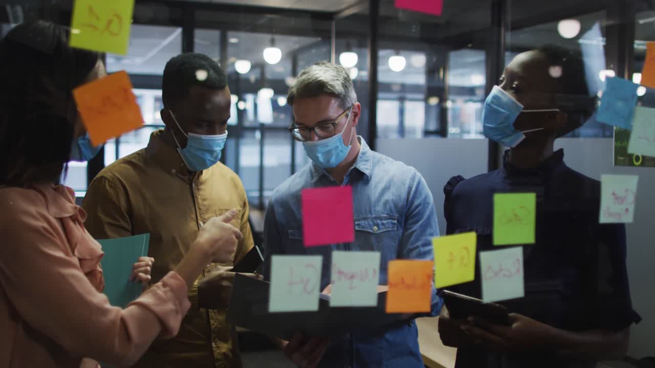 Office colleagues wearing face mask discussing over memo notes on glass board at modern office