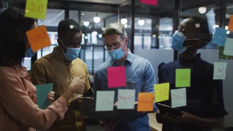 Office-colleagues-wearing-face-mask-discussing-over-memo-notes-on-glass-board-at-modern-office