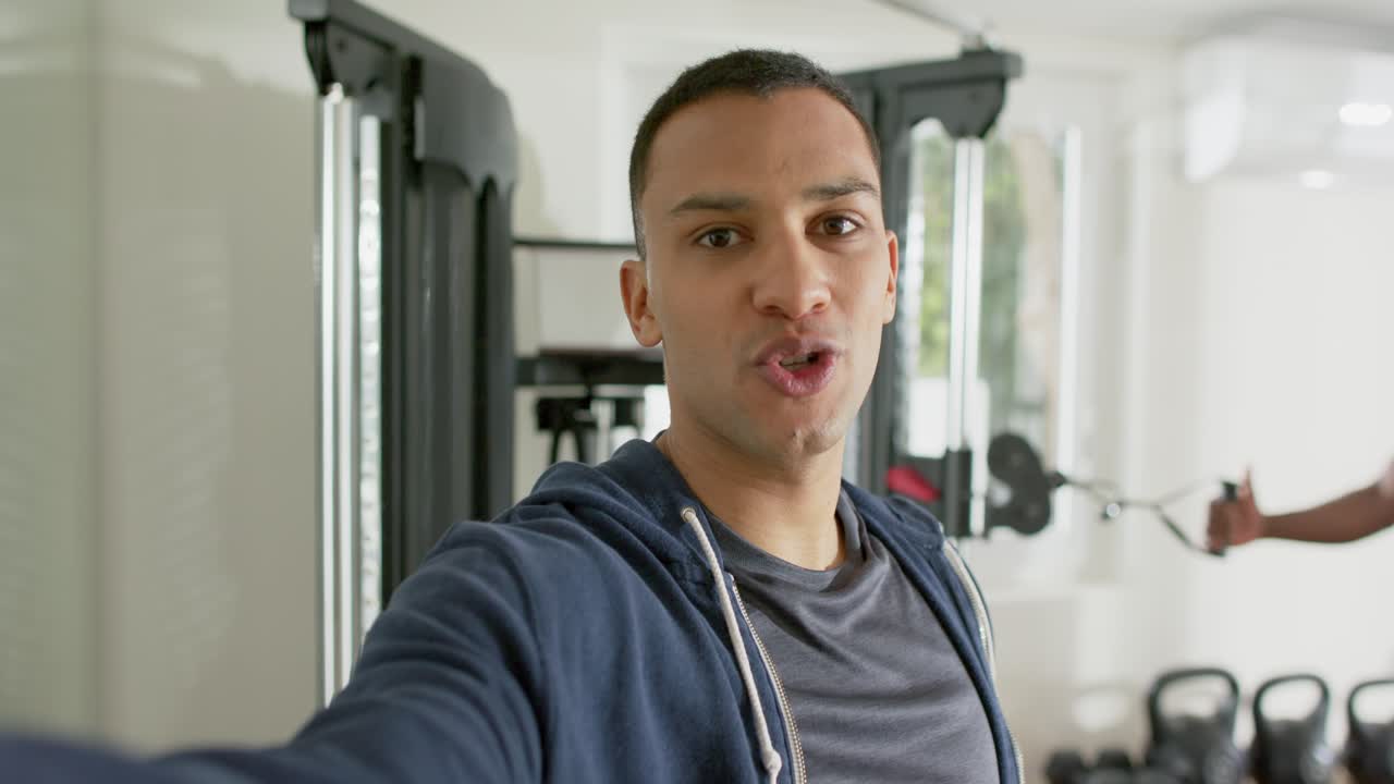 Istruttore ispanico che parla verso la cable machine mentre il partner esegue il rematore con busto flesso nella palestra di casa
