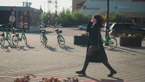 side view of remote worker walking to meeting adjusting hair while carrying handbag, dressed in black coat and boots, urban background features parked cars, green bikes, and blurred glass buildings