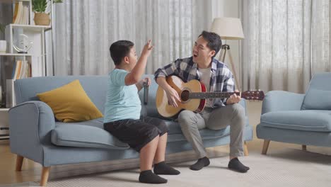 full body of happy asian father and son celebrating for being able to play the guitar and singing together on sofa at home