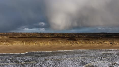 Este-Impresionante-Video-Captura-Una-Cálida-Puesta-De-Sol-Dorada-En-La-Playa,-Con-Nubes-De-Tormenta-En-La-Distancia,-Olas-Rompiendo-Y-Majestuosas-Dunas-De-Arena-Que-Se-Elevan-Sobre-La-Costa