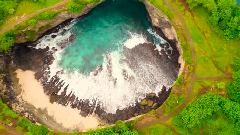 top down aerial drone footage flying further and up from bali broken beach ground cavity that has gushing green ocean sea water waves coming in from a hole in a cliff from the ocean green landscape.