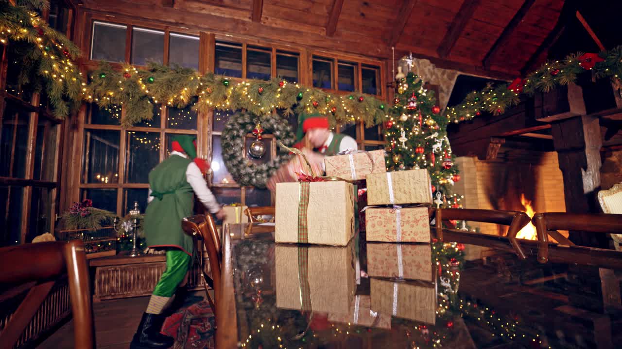 Joyful elves with gift boxes. Happy fairy characters making a pyramid of presents on the table on Christmas decorations background.