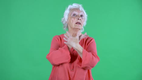 portrait elderly woman looking at the camera with a shocked frightened expression and emotionally gesturing with her hands. gray haired grandmother in red blouse on green screen at studio. close up