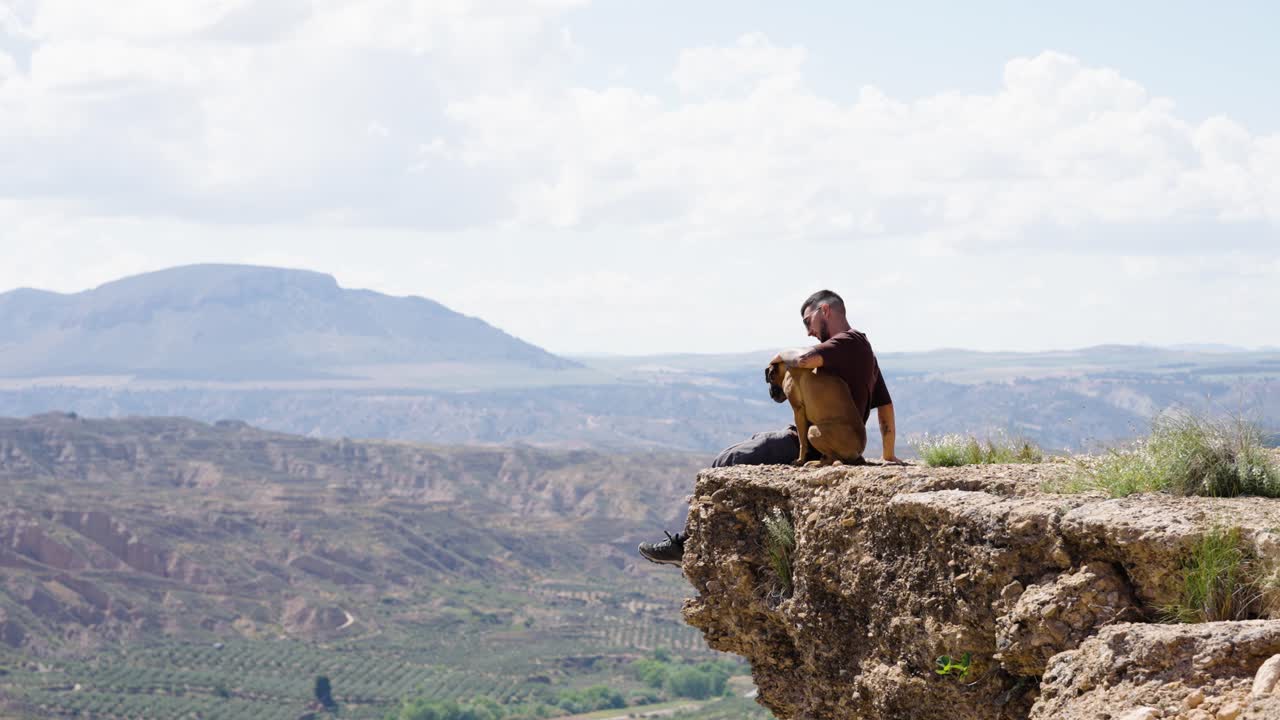 Male hiker with Boxer dog sitting on high cliff above vast valley view