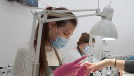 two nail technicians wearing masks and gloves carefully work on client nails under bright task lamps, one filing while the other applies acrylic with precision in a clean, professional salon setting