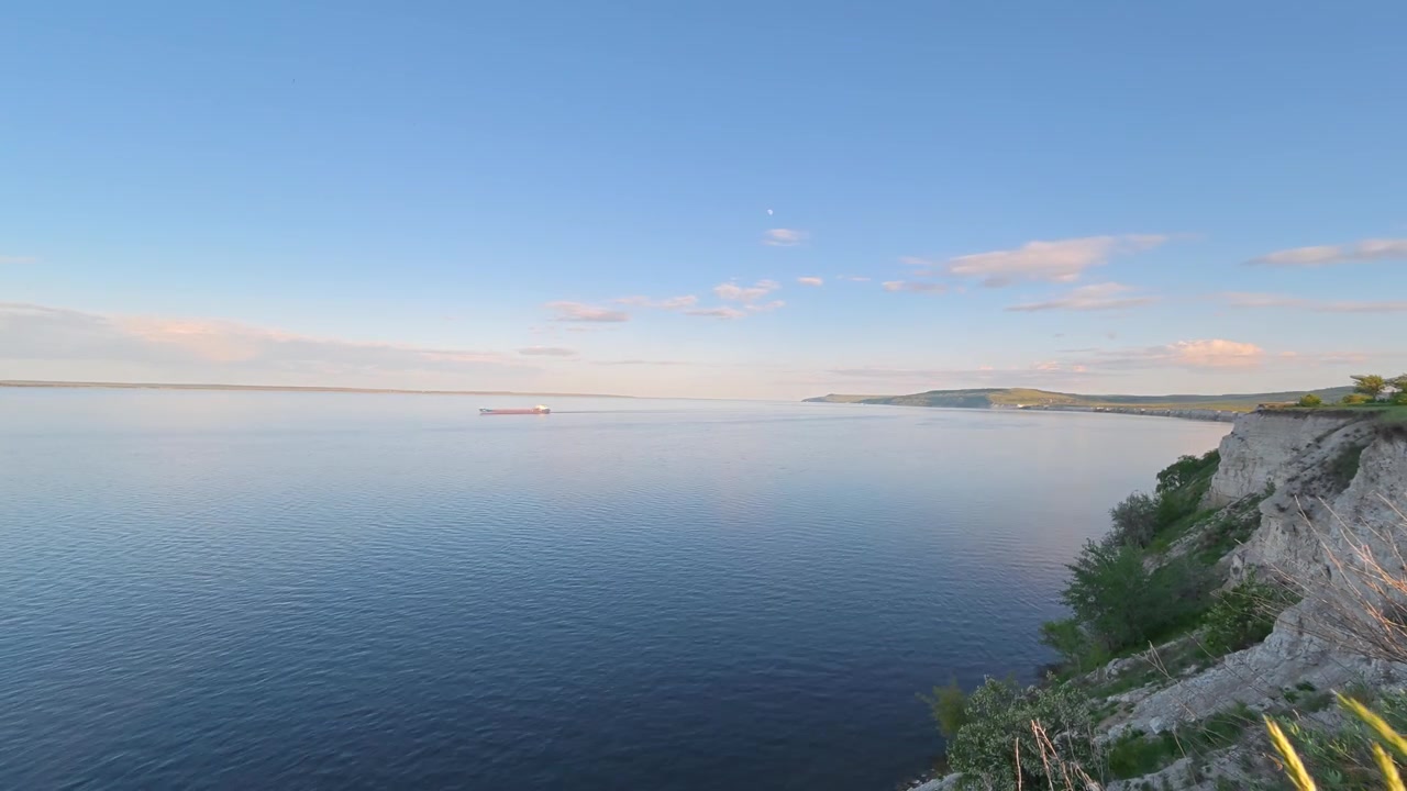 Ein Zeitraffer fängt eine weite Flusslandschaft ein, mit einem Lastkahn, der sich über das ruhige Wasser unter einem klaren blauen Himmel mit vereinzelten Wolken bewegt, von einem hohen Flussufer aus gesehen.