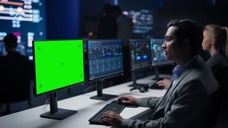 científico de datos masculino confiado trabaja en una computadora de pantalla clave de croma verde en una gran sala de control de infraestructura. los especialistas usan computadoras que muestran gráficos, información.