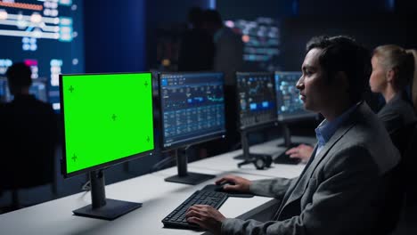 confident male data scientist works on green chroma key screen computer in big infrastructure control room. specialists use computers showing graphs, information. control room professional team