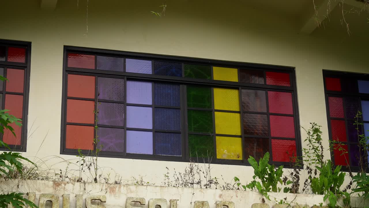 A handheld shot of an old building with large grid window of colorful glass panes in red, blue, yellow, green, and purple, framed by vines and greenery near Mahabang Tanaw, Quezon Province Philippines