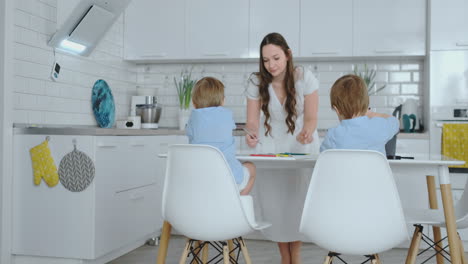 dos niños sentados en la luz de la cocina dibujan lápices dibujos para misiones escolares. mamá mira a los niños y sonríe en la cocina