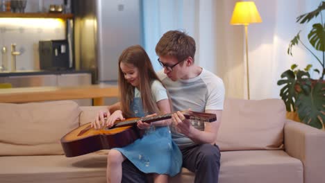 loving father teaching cute little daughter to play guitar sitting together on couch at home