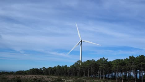 Ferne-Aufnahme-Einer-Einzelnen-Windkraftanlage,-Die-Sich-Dreht,-Mit-Wenigen-Wolken-Dahinter-Und-Einem-Kiefernwald-Darunter