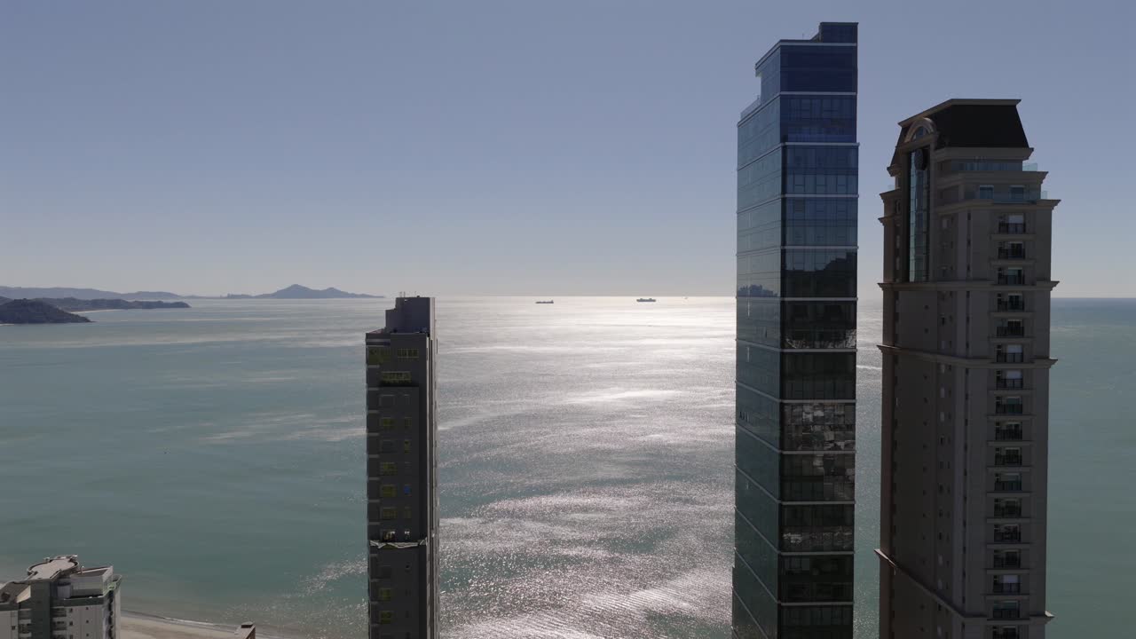 Drone shot flying through and showcasing Balneário Camboriú’s skyline with YachtHouse by Pininfarina, One Tower and Millennium Palace, set against the Atlantic Ocean and distant islands
