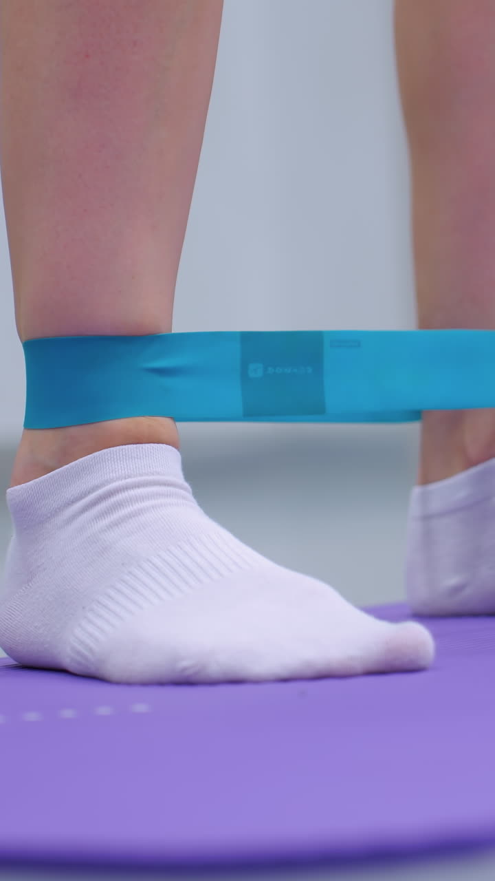 vista de ángulo inferior de primer plano de una persona con calcetines blancos usando una banda de resistencia para estiramientos laterales de las piernas, la alfombra de ejercicio púrpura y la pelota de gimnasia verde en el fondo