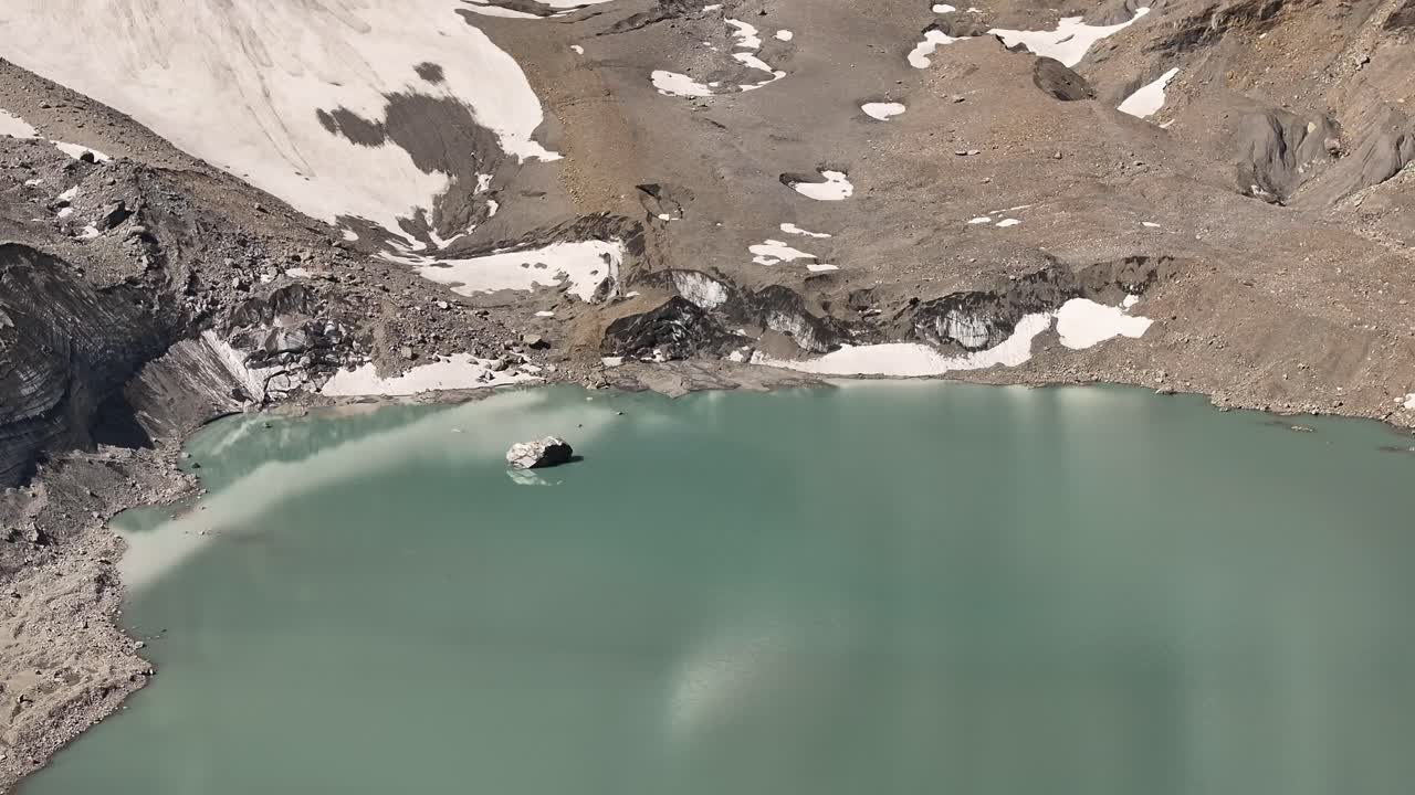 Aerial view of a pale turquoise glacial lake nestled in the mountains of Urnerboden, near the Klausenpass in Kanton Uri, Switzerland. The barren, rocky terrain and patches of snow surrounding the lake