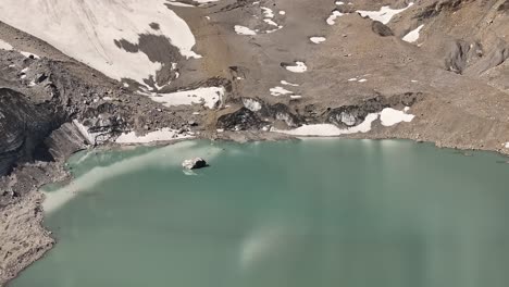 Aerial view of a pale turquoise glacial lake nestled in the mountains of Urnerboden, near the Klausenpass in Kanton Uri, Switzerland. The barren, rocky terrain and patches of snow surrounding the lake