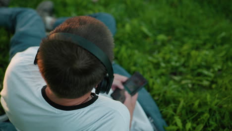 rear view of music producer wearing headphones, nodding his head to the music while operating his phone, he is seated on a grassy field surrounded by vibrant greenery and natural light