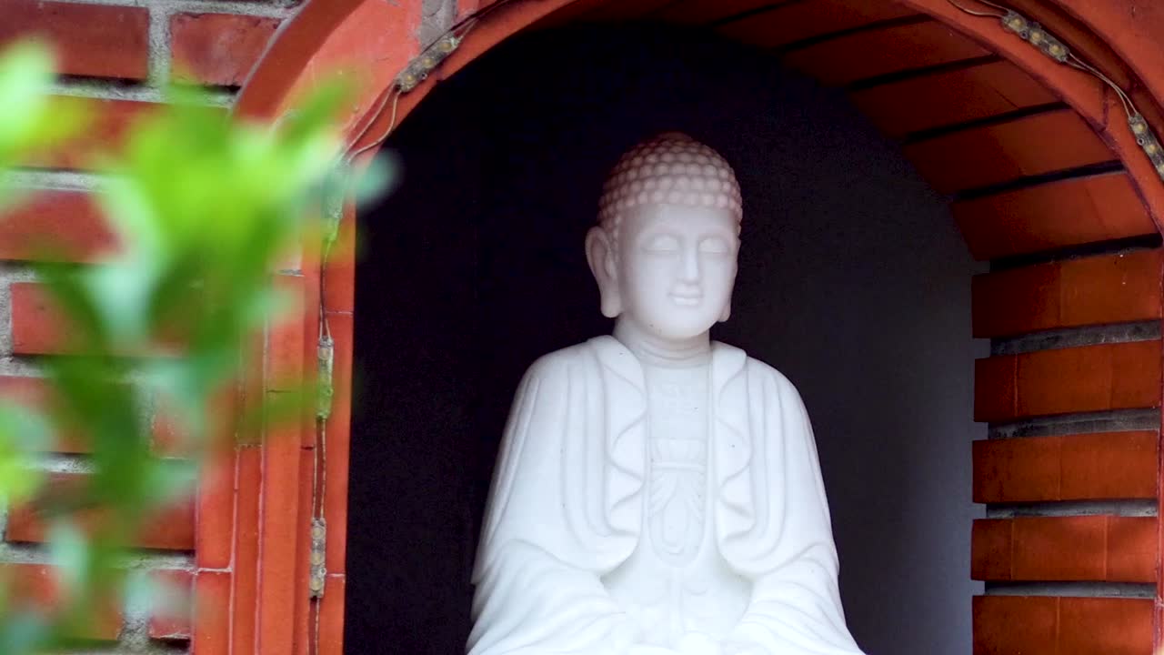 En fredfyldt, hvid Buddha-skulptur står i en murstensniche omkranset af blødt, grønt løv.