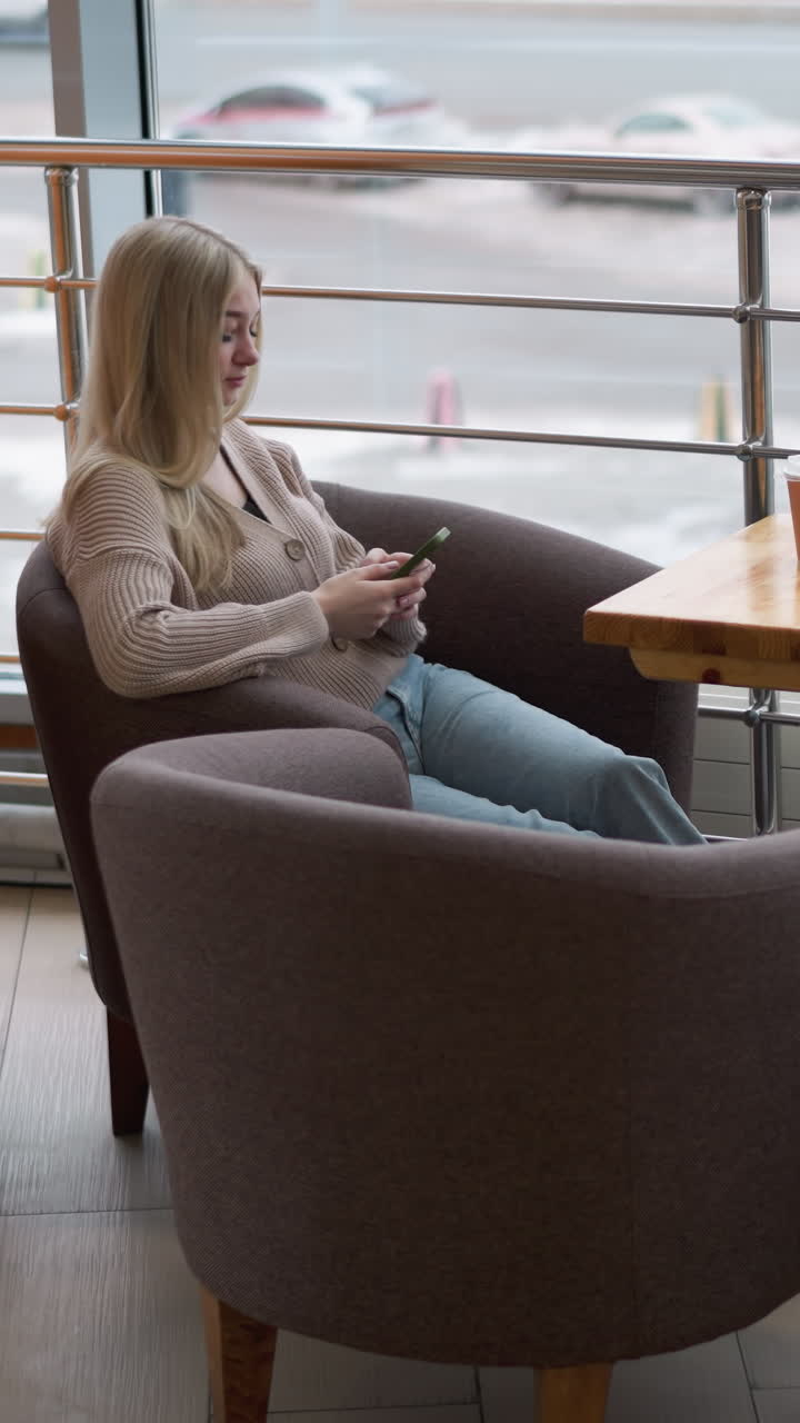 joven sentada sola en el centro comercial con el teléfono en la mano, mirando por la gran ventana con vista a la ciudad ocupada, coches pasando por las calles nevadas mientras disfruta de un momento de paz con café en la mesa de madera