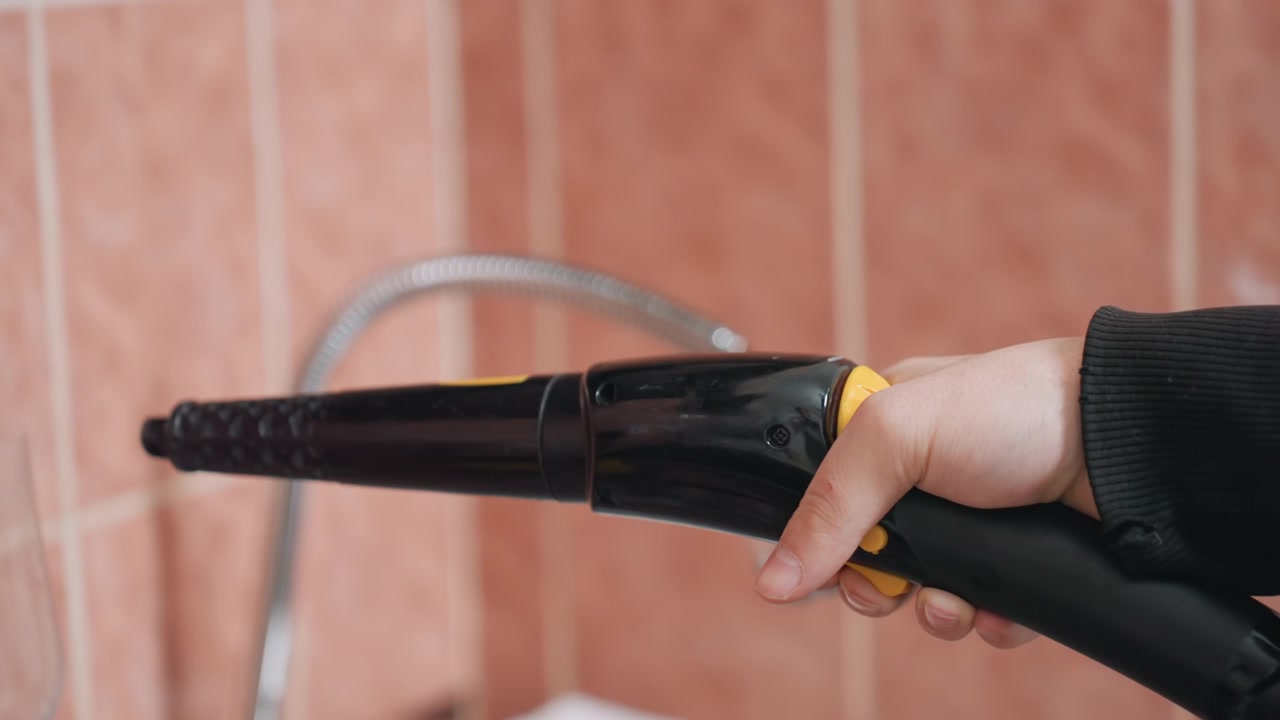 Main en gros plan tenant une buse à vapeur au-dessus de l'évier, par un nettoyeur, prise précise et contrôle de la gâchette utilisés pour le travail de détail des joints et des carreaux, gros plan de l'équipement montrant la poignée, l'embout et le tuyau avec une stabilité.