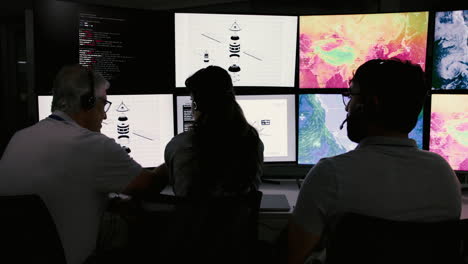 tres personas trabajando en una sala de control con muchas pantallas que muestran datos, mapas y gráficos