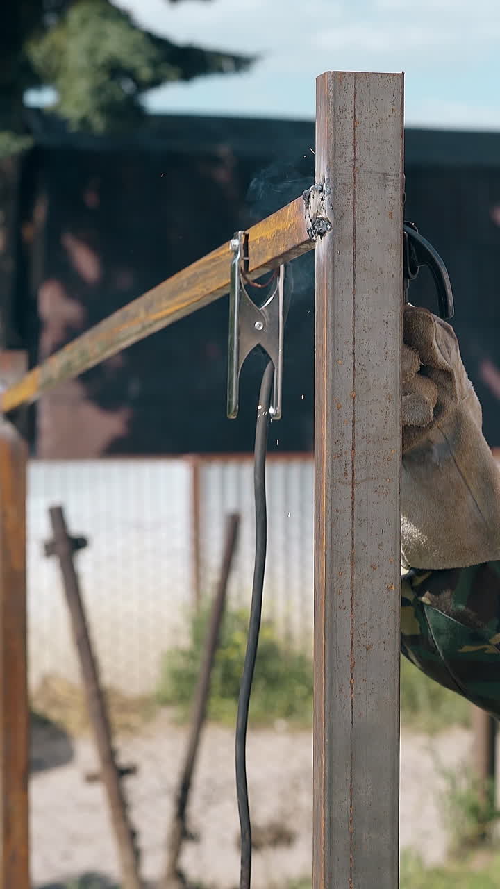Vista laterale di un operaio in abiti da lavoro che fissa le parti superiori di una recinzione metallica con uno strumento di saldatura contro una casa di campagna di giorno.