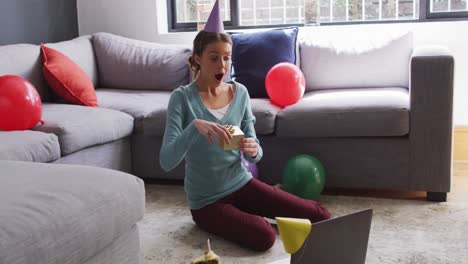 Surprised-woman-in-party-hat-holding-a-gift-box-while-having-a-video-call-on-laptop-at-home