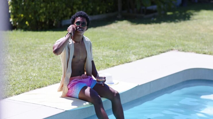 African American man answering call, relaxing on pool edge holding phone and cup wearing sunglasses