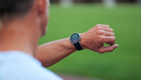 Sportlicher-Männlicher-Läufer,-Der-Auf-Seiner-Smartwatch-Mit-Trainingszeiten-Auf-Der-Grünen-Wiese-Schaut