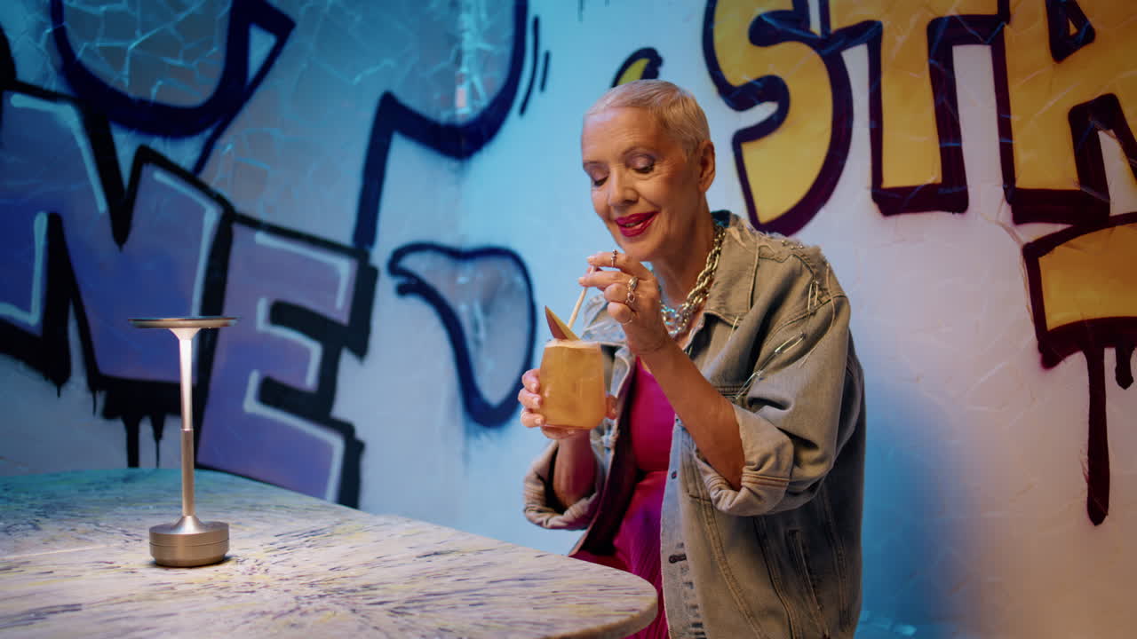 Anziana alla moda che beve un cocktail in un bar con graffiti. Anziana signora allegra in relax.