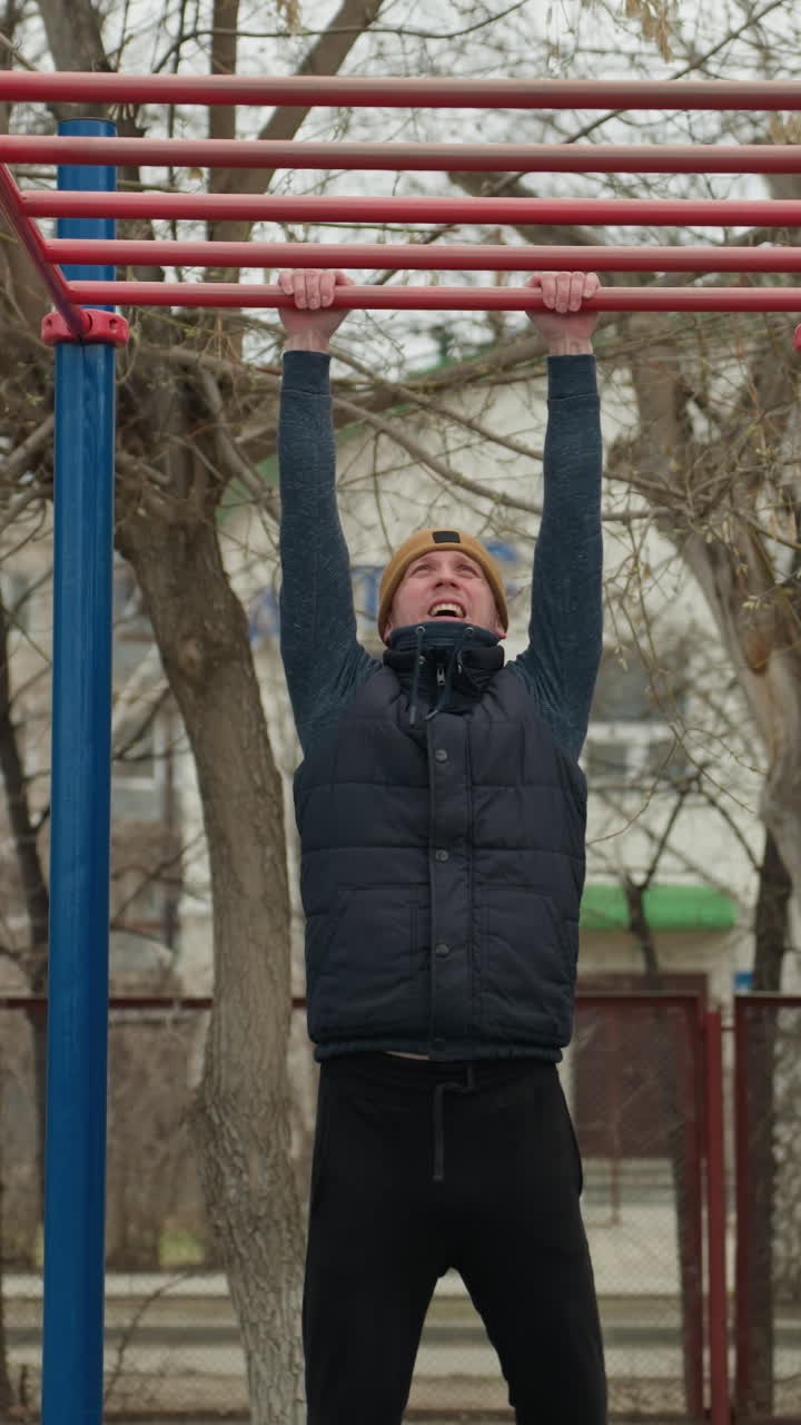 A coach jumps up to grab a red iron bar in an outdoor workout area, with people and building with bare trees in the background