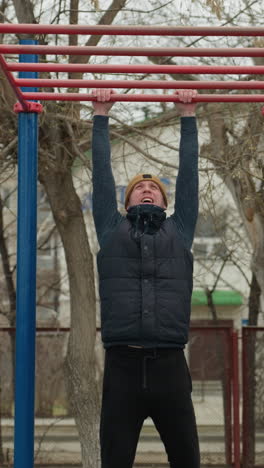 a coach jumps up to grab a red iron bar in an outdoor workout area, with people and building with bare trees in the background