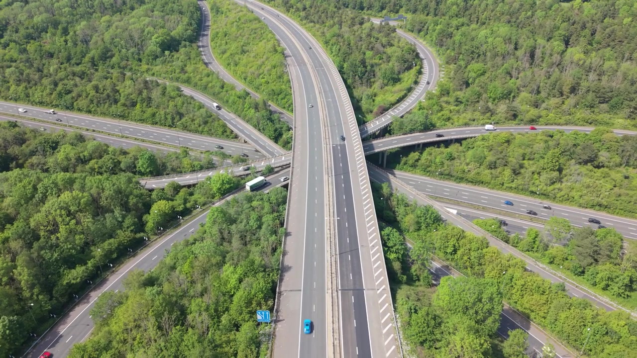High angle view of a vast highway system where multiple roadways merge and cross above lush green trees. Cars and trucks drive on the busy routes during bright daylight.