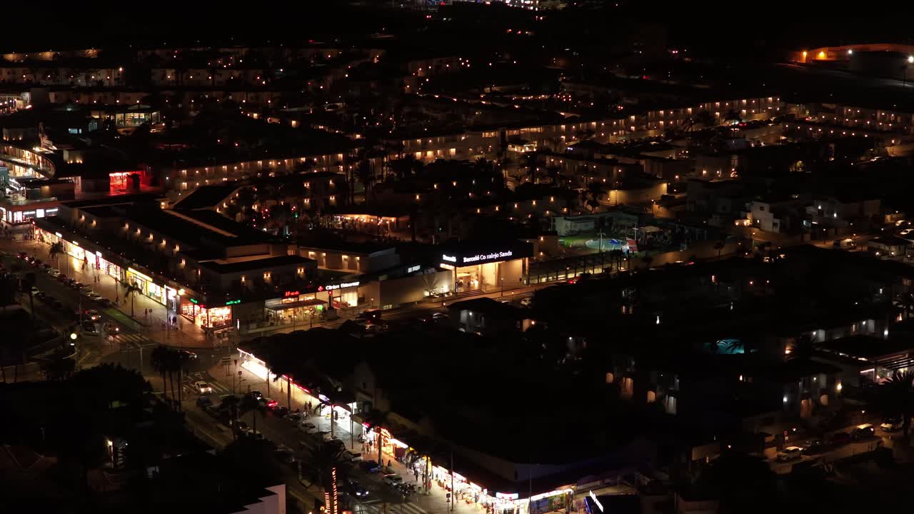 Drone establishing shot over Corralejo Viejo at night reveals Avenida Nuestra Señora del Carmen glowing with storefronts, beach symmetry, and Atlantic dark expanse forming a dramatic coastal contrast