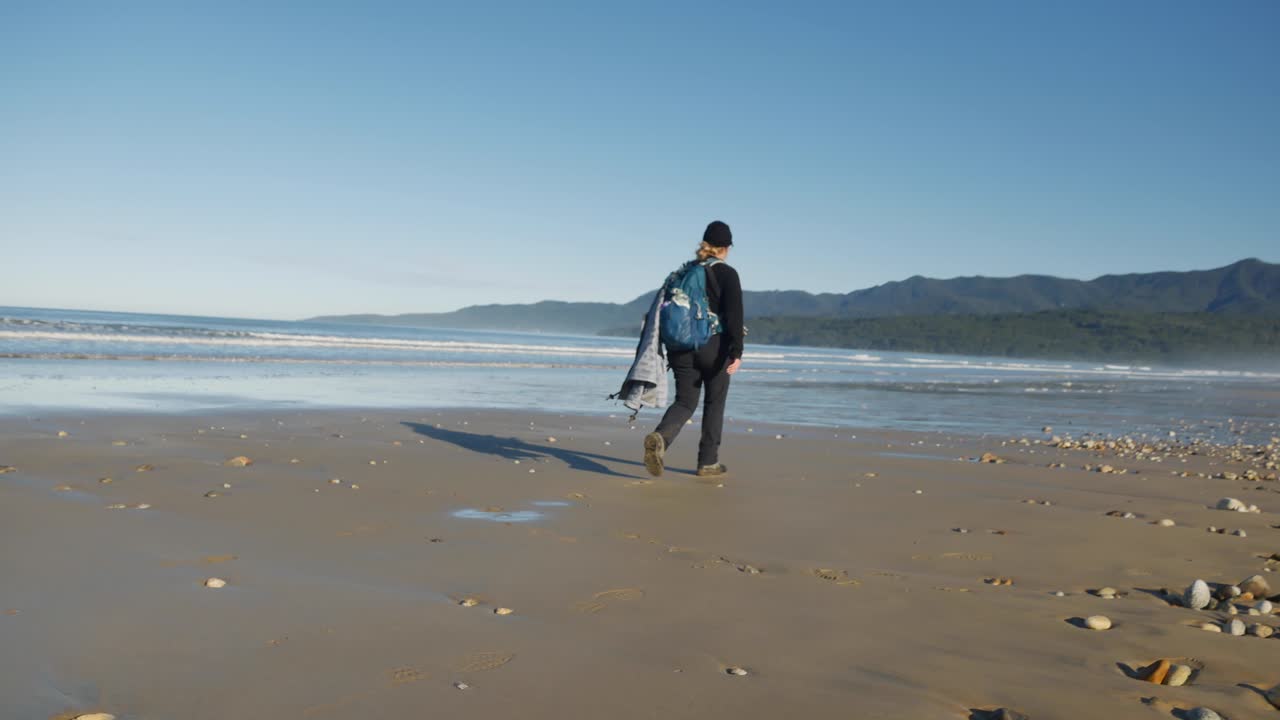 Woman hiker with tripod walks on wide expansive sandy ocean beach