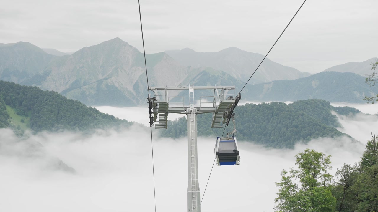 A gondola cabin travels above low cloud with mountain ridges behind it in Gabala in daylight.