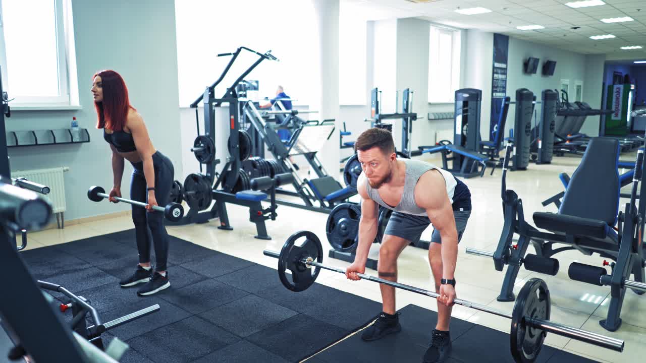 Muscular bearded man and slim woman lifting heavy weights at the gym. Fitness couple in sportswear are doing workout on the background of modern sport center inside.