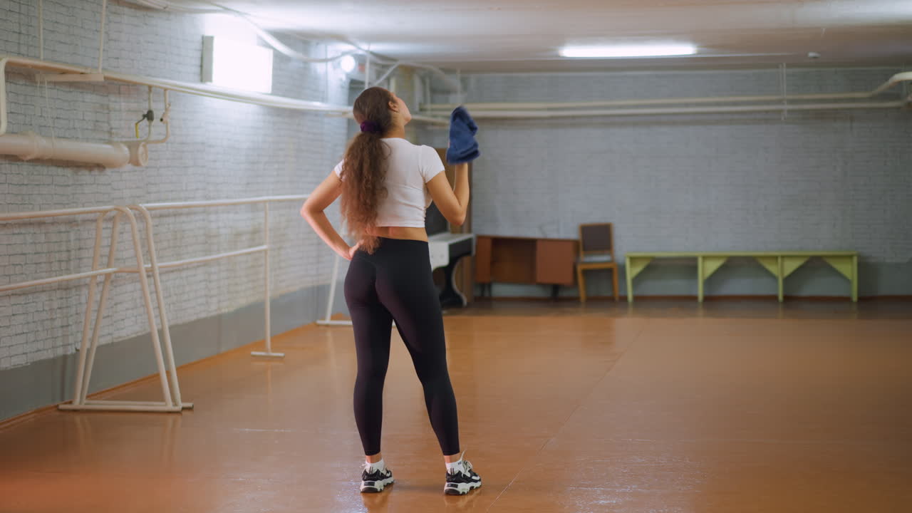 Back view of young woman in white top black leggings and canvas shoes placing hand on waist while wiping sweat from neck with blue towel inside fitness studio after workout showing fatigue