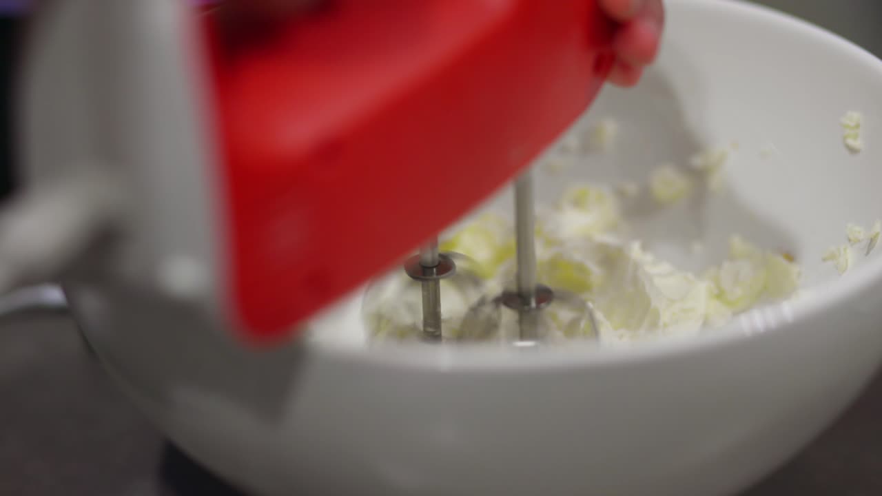 primer plano de una mujer usando una batidora de mano roja para mezclar ingredientes de tarta de queso en un tazón blanco en una cocina